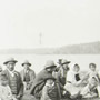 Photograph of a group of Aboriginal men, women and children seated on the ground at a feast, with a lake in the background, Mattagami, July 1906