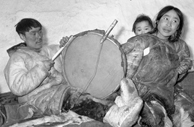 Black-and-white photograph of an Inuit man playing a drum while a woman listens with her baby