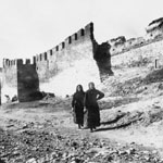 two women walking past a damaged ancient city wall with turrets and guard towers.