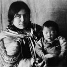 Portrait de Kookooleshook assise sur une chaise avec une toile de fond en arrière-plan, tenant un enfant sur ses genoux, Fullerton, Nunavut, 1904