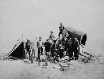 Photographie noir et blanc d'une prairie, avec des gens debout devant une grande charrette et une tente