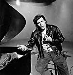 Photograph of Glenn Gould as Theodore Slutz, sitting beside piano, 1980